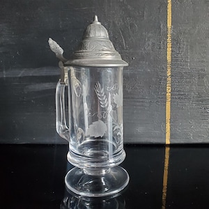 Può includere: Un boccale da birra in vetro trasparente con un disegno inciso di spighe di grano e foglie sul corpo. Il recipiente presenta un coperchio conico in peltro incernierato con incisioni decorative e un robusto manico in vetro con leva in metallo.