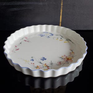 Może przedstawiać: Biała ceramiczna forma do ciasta z falistym brzegiem i kwiatowym wzorem. Naczynie ma delikatny wzór niebieskich, różowych i żółtych kwiatów i liści. Cienka niebieska linia podkreśla krawędź.