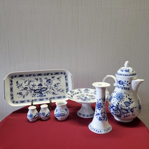 Peut inclure: Ensemble de vaisselle en porcelaine blanche avec des motifs floraux bleus. Comprend un plateau rectangulaire, un présentoir à gâteau, une crémière, une sucrière, un bougeoir et de petits vases. L'ensemble est disposé sur une surface rouge.