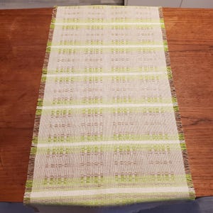May include: A rectangular woven table runner with a beige background and horizontal stripes in shades of green and white. The edges have a fringed detail. The runner is placed on a wooden surface.