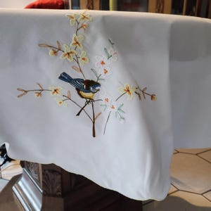Peut inclure: Nappe blanche avec broderie. La broderie représente un oiseau bleu et marron perché sur une branche avec des fleurs jaunes et blanches. La nappe est drapée sur une table en bois.