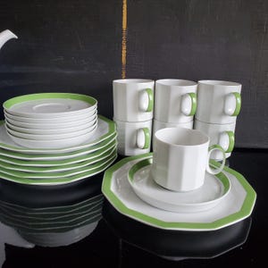 Op de afbeelding: Een witte keramische theeservies met een groene rand. De set bevat kopjes, schotels en borden. De kopjes hebben groene oren. De borden en schotels hebben een groene rand. De set is op een zwart oppervlak geplaatst.
