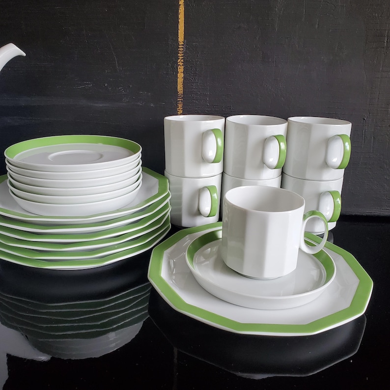 Op de afbeelding: Een witte keramische theeservies met een groene rand. De set bevat kopjes, schotels en borden. De kopjes hebben groene oren. De borden en schotels hebben een groene rand. De set is op een zwart oppervlak geplaatst.