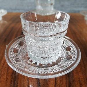 May include: Clear, patterned glass cup and saucer set. The cup sits on a matching saucer, both featuring a detailed, geometric design. The glass is transparent, allowing light to pass through, and is set on a wooden surface.