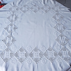 Peut inclure: Nappe blanche avec une bordure brodée. La bordure présente des motifs géométriques et floraux, notamment des losanges et des motifs floraux avec de petits points noirs. La nappe est en tissu blanc doux.