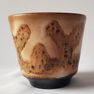 Könnte beinhalten: Eine handgefertigte Keramiktasse mit einem einzigartigen Design. Die Tasse hat eine hellbraune und cremefarbene Außenseite mit dunkelbraunen Sprenkeln und einen schwarzen Boden. Die Tasse hat eine leicht ausgestellte Form und eine glatte, glänzende Oberfläche.