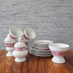May include: A set of white porcelain tableware with a red decorative border. The collection includes stacked cups, plates, a lidded jar, and a pedestal bowl. The items are arranged on a wooden surface against a textured grey background.