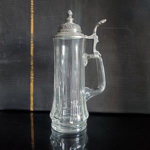 Può includere: Un boccale da birra alto in vetro trasparente con un design a pannelli verticali. Il recipiente include un coperchio incernierato in peltro decorato con un pomello ornamentale e una leva in peltro abbinata fissata a un robusto manico in vetro.