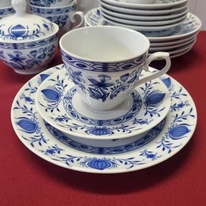 Könnte beinhalten: Weißes Porzellan-Teeservice mit blauem Blumendesign. Das Set umfasst eine Tasse, eine Untertasse und zwei Teller, alle mit passendem blau-weißem Muster. Zusätzliche Tassen und Teller sind im Hintergrund gestapelt.
