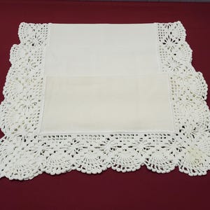 May include: Cream-coloured rectangular doily with a crocheted lace border. The lace features a floral pattern, adding a delicate touch. The doily is set against a deep red background.
