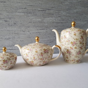 Könnte beinhalten: Ein cremefarbenes Teeservice mit floralem Muster. Das Set besteht aus einer Teekanne, einer Zuckerdose und einem Sahnekännchen. Teekanne und Zuckerdose haben goldene Knäufe, die Teekanne einen goldenen Henkel. Das florale Muster zeigt rosa und gelbe Blüten.