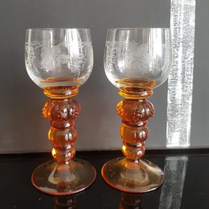 Peut inclure: Deux verres à vin ambrés vintage. Les bols transparents sont gravés d'un motif floral. Les pieds sont d'un ambre plus foncé, avec des formes arrondies et une base circulaire. Les verres sont présentés sur une surface sombre.