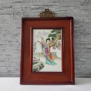 May include: A framed porcelain artwork featuring a woman playing a stringed instrument under a tree. The artwork is set in a dark wood frame with a decorative brass top. The woman is wearing a colorful robe.