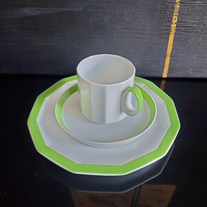 Op de afbeelding: Een witte theekop en schotelset met een heldergroene rand. De theekop heeft een bijpassend groen handvat. De set bevat een groot bord, een kleiner bord en een theekop. De set staat op een zwart oppervlak.