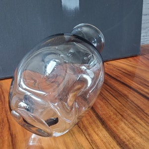 Peut inclure: Un vase en verre transparent de forme ovale, avec une base arrondie et un col étroit. Le vase a une surface lisse et polie et est légèrement incliné, mettant en valeur sa transparence. Le vase est posé sur une surface en bois.