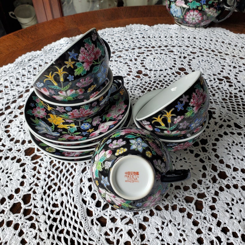 May include: A set of black porcelain teacups and saucers with a floral design. The cups and saucers feature a black background with colorful flowers, leaves, and birds. The cups have black handles and white interiors.