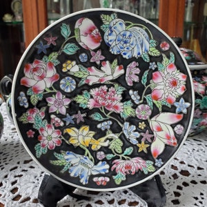 May include: A decorative black plate with a white rim, adorned with a colorful floral design. The flowers include pink, white, blue, and yellow blossoms with green leaves. The plate is displayed on a black stand.