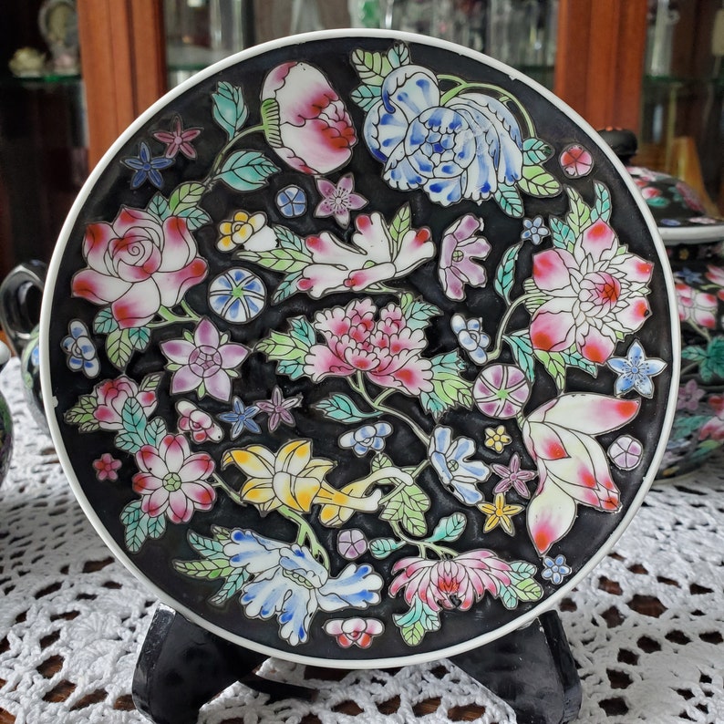 May include: A decorative black plate with a white rim, adorned with a colorful floral design. The flowers include pink, white, blue, and yellow blossoms with green leaves. The plate is displayed on a black stand.