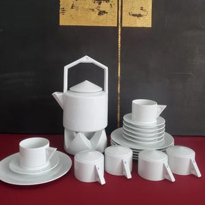 Può includere: Un servizio da tè in porcellana bianca con teiera, tazze, piattini e zuccheriera. La teiera ha un manico geometrico e poggia su un supporto abbinato. Le tazze hanno un design angolare. Il set è su una superficie rossa.
