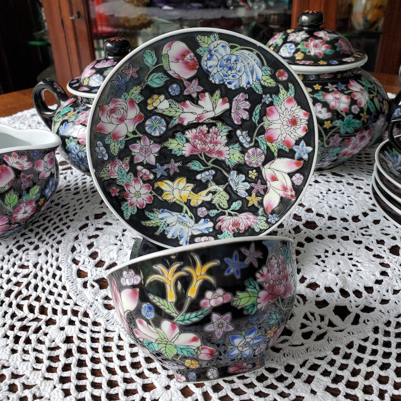 May include: A black porcelain tea set with a floral design. The set includes a teapot, teacups, saucers, and a sugar bowl. The floral pattern features pink, white, yellow, and blue flowers. The set is displayed on a white lace tablecloth.