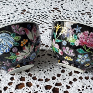 May include: Two black teacups with floral designs. The cups feature colorful flowers in pink, blue, and white, with green leaves. Each cup has a black handle and a white rim. The cups are set on a white lace tablecloth.