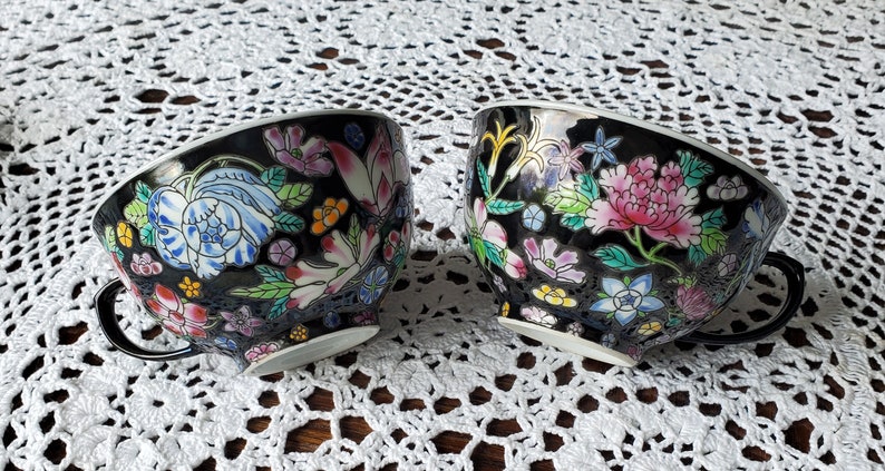 May include: Two black teacups with floral designs. The cups feature colorful flowers in pink, blue, and white, with green leaves. Each cup has a black handle and a white rim. The cups are set on a white lace tablecloth.