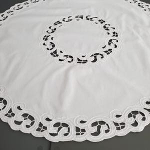 May include: A white, circular tablecloth with detailed cutwork. The border and a central circular motif display a repeating pattern of stylized swirls and shapes, producing a delicate, openwork appearance. The tablecloth is made of a smooth, white fabric.