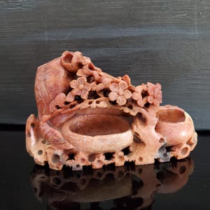 May include: A carved, reddish-brown stone sculpture featuring floral and botanical elements. The sculpture has two bowl-like depressions and intricate details, including flowers and leaves. The piece is set against a dark background.
