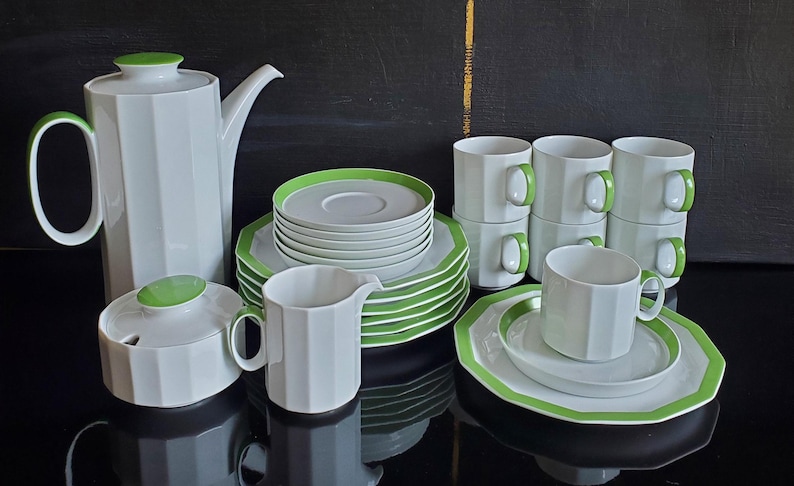 Op de afbeelding: Een wit porseleinen theeservies met een geometrisch ontwerp en groene accenten. De set bevat een theepot, suikerpot, melkkan, kopjes, schotels en borden. De handvatten en randen zijn groen.