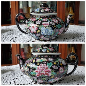 May include: A black ceramic teapot with a lid, featuring a floral design in pink, white, blue, and yellow. The teapot has a black handle and spout, and sits on a white lace tablecloth. The teapot is a decorative item.