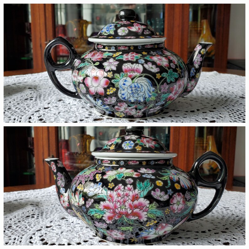 May include: A black ceramic teapot with a lid, featuring a floral design in pink, white, blue, and yellow. The teapot has a black handle and spout, and sits on a white lace tablecloth. The teapot is a decorative item.