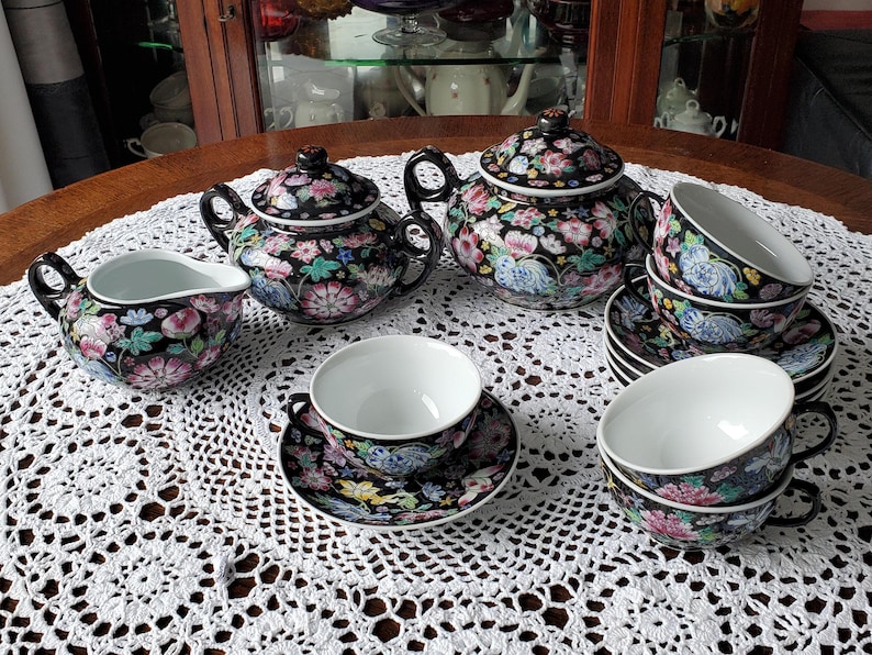 May include: A black tea set with floral designs, including a teapot, creamer, cups, and saucers. The set features pink, blue, and green flowers on a black background. The cups and creamer have white interiors. The set is arranged on a white lace tablecloth.
