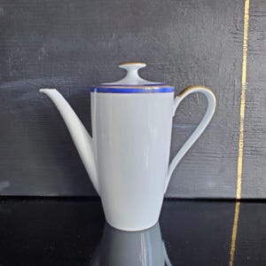 Puede incluir: Cafetera de porcelana blanca con un pico largo y un asa curvada. La cafetera presenta una banda azul alrededor del borde y detalles dorados en el asa y la tapa. La tapa tiene un pequeño adorno dorado. La cafetera está sobre una superficie negra reflectante.
