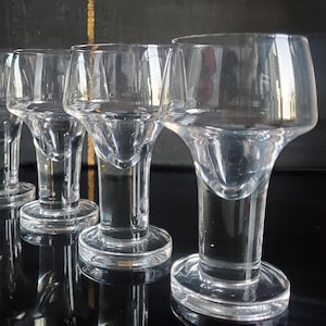 May include: A set of clear, stemmed glasses with a rounded bowl and a short, thick stem. The glasses are arranged in a row, reflecting light and creating a sense of elegance. The base of each glass is circular. The glasses are likely for serving a beverage.