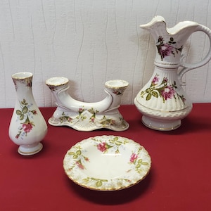 Könnte beinhalten: Eine Sammlung antiker Porzellanartikel: eine Vase, ein doppelter Kerzenhalter, ein Krug und eine kleine Schale. Jedes Stück ist weiß mit einem floralen Rosenmuster in Rosa, Grün und Gold. Die Artikel sind auf einer roten Oberfläche angeordnet.