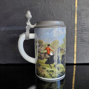 May include: A ceramic beer stein with a hinged lid and a handle. The stein features a detailed illustration of a fox hunt scene with riders on horseback, set against a landscape of trees and a blue sky. The lid and handle hardware are silver-toned.