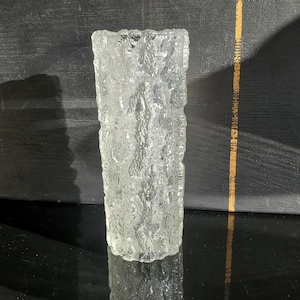 May include: A clear, textured glass vase with a rectangular shape. The vase has a rough, organic surface, and the light reflects off the textured glass. The vase is a decorative item.