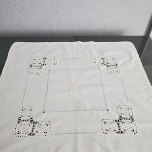 Puede incluir: Mantel rectangular blanco con ribete y esquinas de encaje de ganchillo. El mantel presenta un diseño bordado central con las iniciales "MK" y un borde cuadrado. Las esquinas tienen diseños florales decorativos.