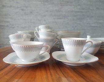 May include: A white porcelain tea set with a light blue geometric design. The set includes teacups, saucers, a sugar bowl, and plates. The cups and saucers have gold trim. The tea set is arranged on a wooden surface.