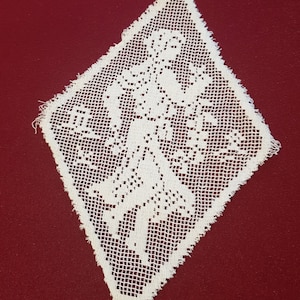 Puede incluir: Un tapete de encaje blanco en forma de diamante con un diseño floral. El delicado patrón calado se destaca sobre un fondo rojo oscuro, mostrando los intrincados detalles del textil hecho a mano. Los bordes están ligeramente deshilachados.