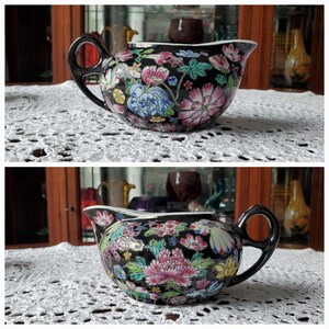 May include: A black ceramic creamer with a floral design featuring pink, blue, and yellow flowers. The creamer has a black handle and spout. The creamer is sitting on a white, lace tablecloth.