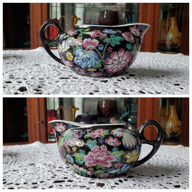 May include: A black ceramic creamer with a floral design featuring pink, blue, and yellow flowers. The creamer has a black handle and spout. The creamer is sitting on a white, lace tablecloth.