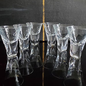 Puede incluir: Seis vasos de chupito transparentes con un acabado texturizado y un diseño angular. Los vasos están dispuestos en fila, reflejándose en una superficie oscura y brillante. El fondo es una superficie negra texturizada con una raya dorada.