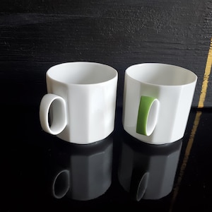 Op de afbeelding: Twee witte keramische mokken met een geometrisch ontwerp. E&eacute;n mok heeft een witte handgreep en de andere een groene handgreep. De mokken staan op een reflecterend zwart oppervlak.
