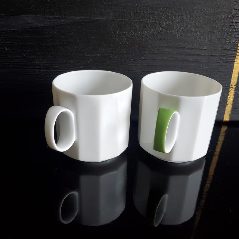 Op de afbeelding: Twee witte keramische mokken met een geometrisch ontwerp. E&eacute;n mok heeft een witte handgreep en de andere een groene handgreep. De mokken staan op een reflecterend zwart oppervlak.