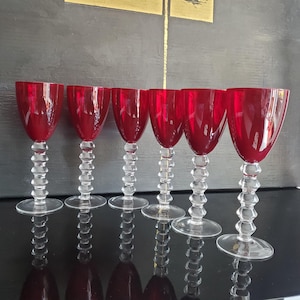 May include: A set of six wine glasses featuring deep red bowls and clear glass stems with a stacked, spherical bead design. Each glass has a wide, circular base and a smooth, rounded rim, creating a uniform appearance across the entire collection.