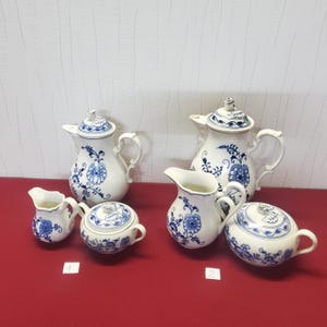 Può includere: Una collezione di pezzi da portata in ceramica bianca con disegni floreali blu. Il set comprende due teiere con coperchi, due lattine e due zuccheriere coperte. I pezzi sono disposti su una superficie rossa.