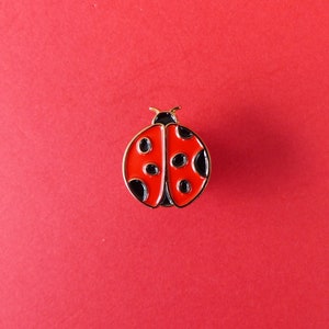 May include: A red and black ladybug pin with gold accents. The ladybug has black spots and a gold outline.