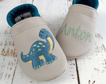 May include: A pair of gray leather baby shoes with a dinosaur design. One shoe has the name Anton embroidered on it. The shoes are made by Little Rabbit.