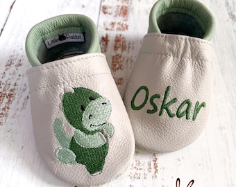 May include: A pair of light gray leather baby slippers with mint green trim. One slipper features an embroidered green dinosaur, while the other displays the name Oskar in green embroidery. A Little Rabbit brand label is visible inside.